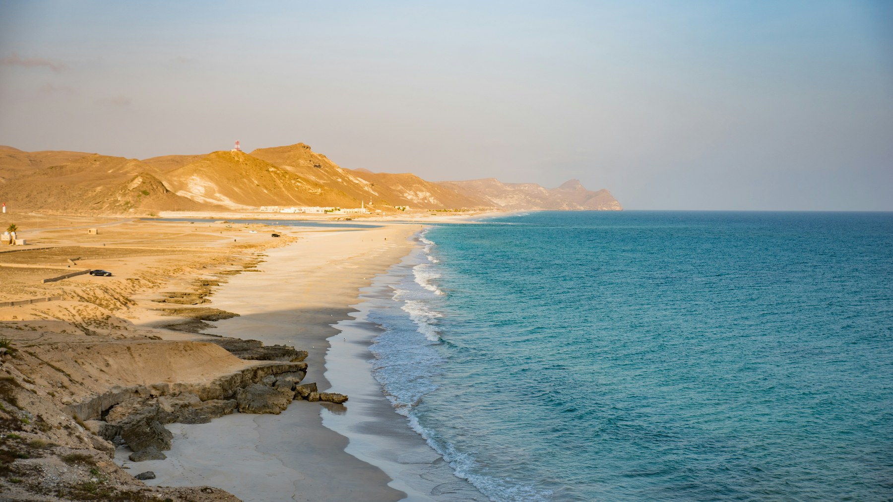 A scenic view of a sandy beach meeting the turquoise ocean, with rocky hills in the background under a clear sky.